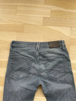 Grå jeans från Jack & Jones - Snygga grå jeans från Jack & Jones med klassisk femficksdesign och normal passform. Jeansen har en lätt tvättad look och detaljer i silverfärg på knappar och nitar. Perfekta för en avslappnad och trendig stil.