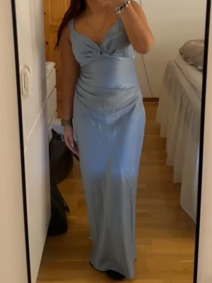 Ljusblå långklänning från Adoore - Säljer en elegant ljusblå långklänning från Adoore med smala axelband och v-ringad urringning. Klänningen har en figurnära passform och är tillverkad i ett mjukt, glansigt material som ger en lyxig känsla. Perfekt för speciella tillfällen.