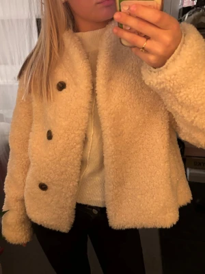 Kräm färgad teddyjacka med knappar - Mysig teddyjacka med knappar framtill. Jackan har en boxy passform, lång ärm och fluffigt material som ger en trendig look. Perfekt för att hålla värmen under kyliga dagar.