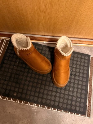 Bruna fluffiga tofflor med foder - Mysiga bruna tofflor med vitt fluffigt foder och dekorativ söm runt öppningen. Tofflorna har en högre ankelmodell och är gjorda i mockaimitation med mjuk sula. Perfekta för att hålla fötterna varma hemma.