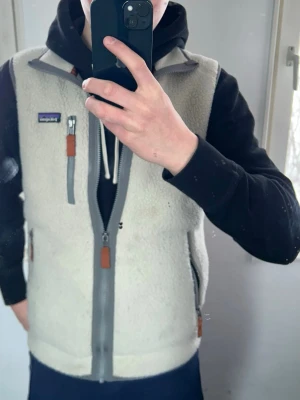 Beige fleeceväst från Patagonia - Säljer en beige fleeceväst från Patagonia med grå dragkedjor och bruna detaljer. Västen har två sidofickor med dragkedja och en bröstficka med logga. Jag säljer den för att jag vill köpa en ny i en annan färg. Den har storlek S och passar perfekt på mig och jag är 175 cm lång. Den har inga slitningar eller hål alls. Bra skick.
