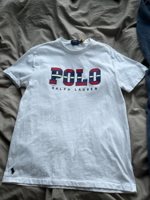 Vit POLO t-shirt från Ralph Lauren - Snygg vit t-shirt från Ralph Lauren med stort POLO-tryck i rött, blått och vitt på bröstet. Klassisk passform med korta ärmar och rund hals. Liten broderad logga nere vid fållen. Tillverkad i mjuk bomull för skön känsla.