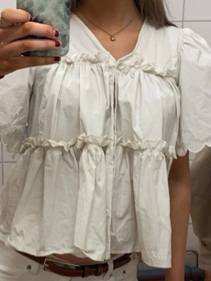 Vit puffärmad blus med volanger - Söt vit blus med puffiga korta ärmar och volangdetaljer. Blusen har en lös passform, knappar framtill och markerad midja med rynk. Perfekt för en trendig och avslappnad look. 