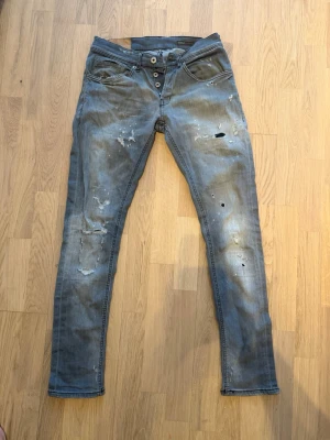 Grå slitna jeans med knappar - Säljer ett par grå jeans med skinny fit och slitningar samt hål på framsidan. Jeansen har knappgylf och fem fickor. Snyggt tvättade detaljer och en avslappnad streetstil. Perfekta för dig som gillar en tuff look.