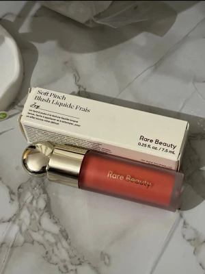 Rare Beauty Soft Pinch Liquid Blush - Flytande rouge från Rare Beauty i nyansen Joy. Kommer i en frostad glasflaska med guldfärgat lock och applikator. Ger en fräsch, persikoorange ton till kinderna och är lätt att blanda ut för ett naturligt resultat.