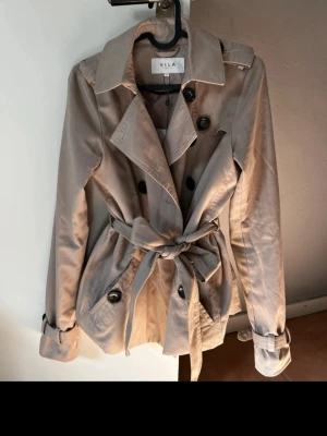 Beige trenchcoat från VILA - Stilren beige trenchcoat från VILA med dubbelknäppning, klassisk krage och knytskärp i midjan. Jackan har axelklaffar, fickor med knapp och justerbara ärmslut. Perfekt för dig som vill ha en tidlös och trendig look. Helt ny med prislapp, köpt för 500kr. Perfekt trenchcoat till våren! 🥰