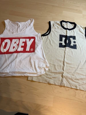 Vita linnetoppar från OBEY & DC - Två snygga linnetoppar, en från OBEY med stor röd logga framtill och en från DC med svart logga och svarta kontrastkanter. Båda är ärmlösa och har avslappnad passform, perfekta till sommaren eller streetwear-stilen.