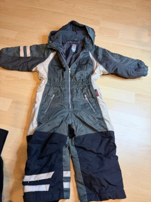 Grå täckbyxa med huva från New - Täckbyxa i grått och svart med beige detaljer från New Generation. Har huva, dragkedja framtill, två fickor med dragkedja och reflexdetaljer på benen. Fodrad insida för extra värme, perfekt för kalla dagar utomhus.