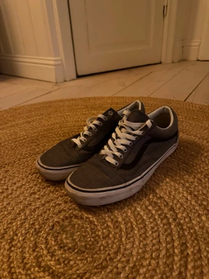 Gråa Vans Old Skool sneakers - Säljer ett par klassiska Vans Old Skool sneakers i grått tyg med vita detaljer och vit sula. Skorna har den ikoniska svarta Vans-sidan och vita skosnören. Perfekta för dig som gillar en clean och tidlös stil.