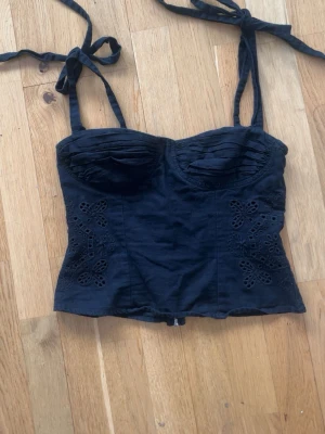 Svart topp med knytband - Svart topp med knytbara axelband och dekorativa broderade detaljer på sidorna. Toppen har en dragkedja därbak.Toppen har ett litet hål precis vid dragkedjan, som du ser på bild 4. Påverkar dock inte själva dragkedjan. Toppen är i storlek S.