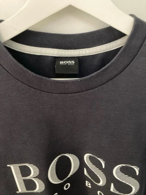 Mörkblå sweatshirt från Hugo Boss - Säljer en mörkblå sweatshirt från Hugo Boss med stort vitt logotryck på bröstet. Tröjan har rund halsringning, långa ärmar och ribbade muddar vid ärmslut och nederkant. Tillverkad i mjuk bomull för skön känsla.