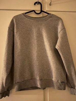 Grå basic sweatshirt utan huva - En enkel och stilren grå sweatshirt i mjukt bomullsmaterial. Tröjan har rund halsringning, ribbade muddar vid ärmslut och nederkant samt en avslappnad passform. Perfekt för en chill och casual look.