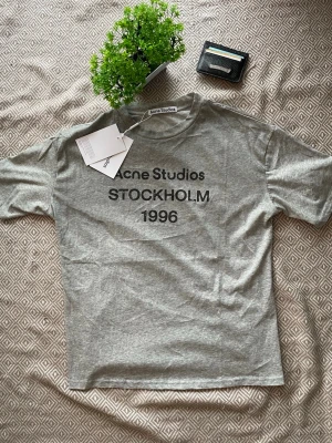 Grå Acne Studios t-shirt Stockholm - Tja! Jag säljer en Snygg grå t-shirt från Acne Studios med tryck 'STOCKHOLM 1996' på bröstet. Klassisk passform och rund hals. Perfekt basplagg med stilren design och mjukt bomullsmaterial.