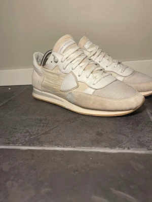 Vita Philippe Model sneakers - Snygga vita sneakers från Philippe Model med klassisk låg siluett. Skorna har ovandel i textil och mocka, vita snören och diskret logga på sidan. Yttersulan är i gummi med beige detaljer. Perfekta för en clean och stilren look.