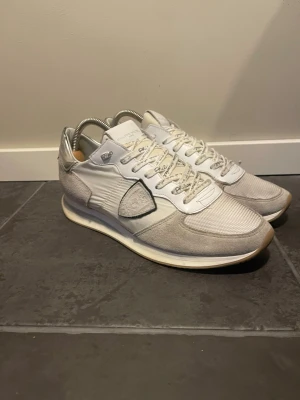 Vita sneakers från Philippe Model - Stilrena vita sneakers från Philippe Model med detaljer i mocka och mesh. Skorna har en låg silhuett, vita snören med text och en beige sula. Klassisk logotyp på sidan och silverfärgade detaljer vid hälen. Perfekta för en clean och modern look.