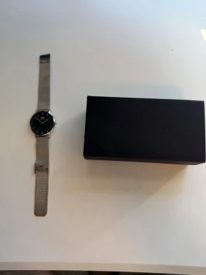 Silverklocka från Daniel Wellington - Stilren klocka från Daniel Wellington med svart urtavla och mesh-armband i silverfärgat metall. Minimalistisk design med DW-logga på urtavlan och justerbart spänne. Perfekt accessoar för dig som gillar klassisk och modern stil.