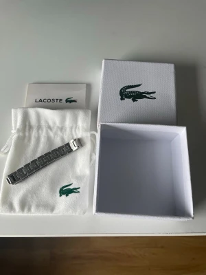 Silverfärgat armband från Lacoste - Snyggt silverfärgat armband från Lacoste i rostfritt stål. Armbandet har en stilren, rektangulär länkdesign med diskret Lacoste-logga på vissa länkar. Kommer med originalpåse och box med den klassiska gröna krokodilen.