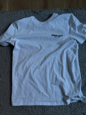 Vit Essentials Fear of God t-shirt - Säljer en vit t-shirt från Essentials Fear of God med tryck på bröstet och ryggen. Klassisk rund hals och korta ärmar. Tillverkad i mjuk bomull för en clean och enkel look. Perfekt för dig som gillar streetwear och minimalistisk stil.