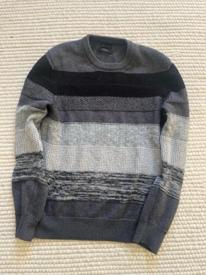Randig stickad tröja från Diesel - Snygg stickad tröja från Diesel med breda horisontella ränder i olika nyanser av grått och svart. Tröjan har rund halsringning och ribbade muddar. Perfekt för dig som gillar en stilren och modern look.
