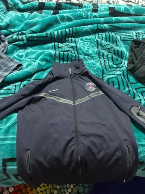 Mörkblå PSG Nike träningsjacka - Snygg mörkblå träningsjacka från Nike med PSG-logga på bröstet och vit Nike-swoosh. Jackan har hel dragkedja framtill, två fickor med dragkedja och en grå detalj över bröstet. Perfekt för fotbollsträning eller chill.