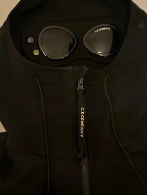Svart vindjacka från C.P. Company - Säljer en svart vindjacka från C.P. Company med ikoniska inbyggda goggles i huvan. Jackan har dragkedja framtill, snörning vid huvan och en clean, modern look. Perfekt för dig som gillar streetwear och vill sticka ut med detaljer.