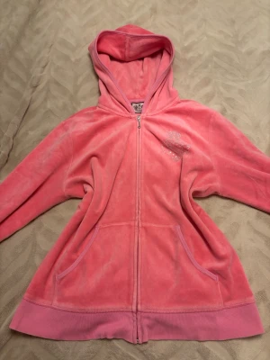 Rosa velourhoodie från Juicy Couture - Säljer en klassisk rosa velourhoodie från Juicy Couture med dragkedja framtill, huva och fickor. På bröstet finns en liten logga med strass och på ryggen ett stort emblem med dekorativa detaljer. Perfekt för dig som gillar Y2K-stil och mjuka material.