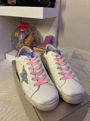 Golden goose  - Snygga vita sneakers från Golden Goose med rosa snören, silverglittrig stjärna på sidan och blå logga på plösen. Skorna har handskriven text på sulan och beige detalj vid hälen. Perfekta för dig som vill sticka ut med coola detaljer.