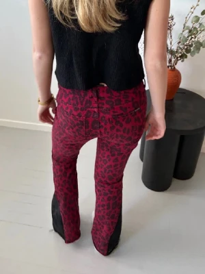 Röda leopardmönstrade jeans - Säljer ett par röda jeans med svart leopardmönster och svarta detaljer längst ner på benen. Jeansen har klassisk femficksmodell och omsydda bootcut. Perfekta för dig som vill sticka ut med en cool och trendig look. (Lågmidjade), modellen är 168