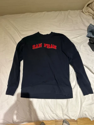 Mörkblå sweatshirt Jack & Jones - Mörkblå sweatshirt från Jack & Jones med röd text 'SAN FRAN' på bröstet. Klassisk rund halsringning och ribbade muddar vid ärmslut och nederkant. Perfekt för en avslappnad och cool stil.