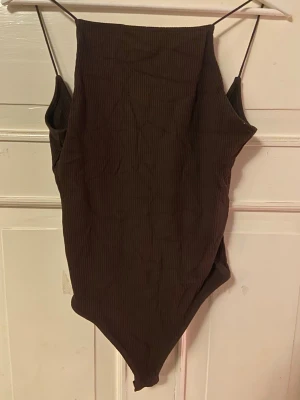 Brun ribbad body med smala axelband - Tittar intresset på en mörkbrun ribbad body med smala axelband och djup urringning bak. Materialet är stretchigt och följsamt, perfekt för att styla med jeans eller kjol. Storlek M men passar även S/XS. Pris går att diskutera.