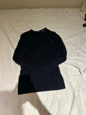 Mörkblå stickad tröja Tommy Hilfiger - Snygg mörkblå stickad tröja från Tommy Hilfiger med rund halsringning och diskret logga på bröstet. Tröjan har långa ärmar och ribbade muddar vid ärmslut och nederkant. Perfekt för dig som gillar klassisk och stilren design.