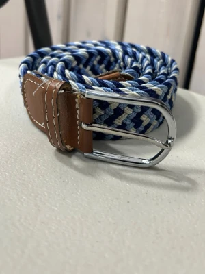 Randigt blått och vitt flätat bälte - Snyggt flätat bälte i blått och vitt med bruna skinn-detaljer och silverfärgat spänne. Perfekt accessoar för att lyfta din outfit med en cool och avslappnad vibe. Passar till jeans eller shorts och ger en fräsch touch.