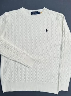 Vit kabelstickad tröja Polo Ralph Lauren - Kabelstickad vit tröja från Polo Ralph Lauren med klassisk rund halsringning och den ikoniska marinblå logotypen broderad på bröstet. Tröjan har långa ärmar och ribbade muddar vid ärmslut och nederkant. Perfekt för en clean och stilren look. Fråga för mer bilder!