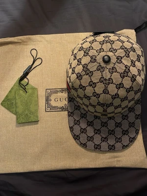 Grå  GG Supreme keps från Gucci - Säljer en grå keps från Gucci med det klassiska GG Supreme-mönstret i brunt. Kepsen har böjd skärm, svart knapp på toppen och justerbart band bak. Tillverkad i canvasmaterial och har en diskret röd-grön detalj på sidan. Perfekt för dig som gillar exklusiva accessoarer. 