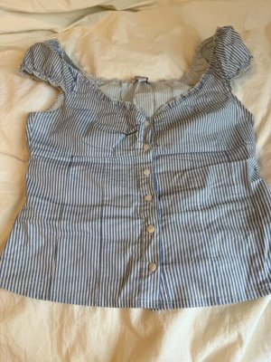 Blårandig blus från Subdude - Söt blårandig blus från Bahaed med knappar framtill och volangdetaljer vid axlarna. Blusen har en v-ringad urringning och är ärmlös, perfekt för varma dagar. Tillverkad i ett lätt bomullstyg som känns luftigt och skönt.