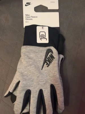 Nike Club Fleece grå handskar - Nike Club Fleece Gloves i grått med svart Nike-logga. Handskarna har ribbade muddar och är tillverkade i mjukt fleece-material. Perfekta för kyliga dagar och har touch-funktion på pekfingret.