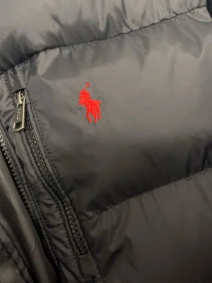 Ralph lauren jacka  - Säljer en svart pufferjacka från Polo Ralph Lauren med röd broderad logga på bröstet. Jackan har huva, två dragkedjeförsedda fickor och justerbar nederkant med snören. Perfekt för kalla vinterdagar och riktigt snygg streetstil.