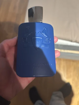 Parfums de Marly parfym blå flaska - Snygg parfymflaska från Parfums de Marly i matt blått med silverfärgat lock. Flaskan har en rektangulär form med rundade kanter och dekoreras av två upphöjda hästar och en sköld på framsidan. Perfekt för dig som gillar exklusiva dofter.  INGEN PARFYM I FLASKAN 