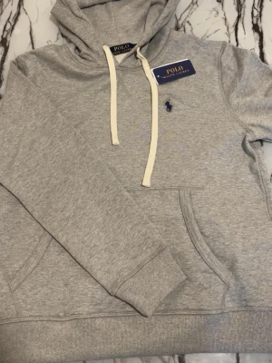 Ralph lauren hoodie  - Snygg grå hoodie från Polo Ralph Lauren med klassisk huva och vita dragsnören. Broderad marinblå logga på bröstet och stor magficka framtill. Tillverkad i mjuk bomullsblandning som känns skön mot huden. Perfekt för en avslappnad och stilren look. 