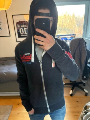 Mörkgrå Superdry zip hoodie - Säljer en mörkgrå zip hoodie från Superdry med tryck i rött och blått på bröstet och ärmen. Hoodien har vita dragsnören, hel dragkedja och mjukt material. Perfekt för dig som gillar streetwear och vill ha en snygg och bekväm hoodie.