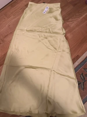 Ljusgul satinkjol från Lindex  - Snygg ljusgul långkjol i satin från Lindex . Kjolen har en slät och glansig yta med enkel design och rak passform. Perfekt för dig som gillar stilrena och trendiga plagg med en mjuk känsla.