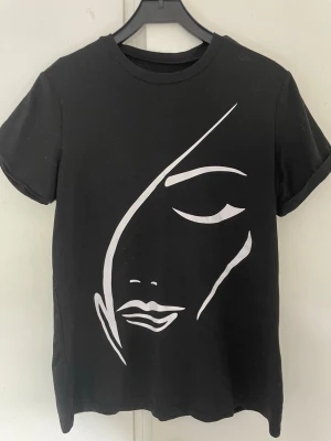 Svart t-shirt med vit ansiktsgrafik - Svart t-shirt i bomull med en stor vit grafisk illustration av ett ansikte på framsidan. T-shirten har rund hals, korta ärmar med uppvik och en normal passform. Perfekt för dig som gillar stilrena och konstnärliga prints.