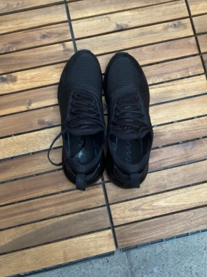 Svarta Nike Air Max 270 sneakers - Säljer ett par helsvarta Nike Air Max 270 sneakers med mesh-överdel och synlig Air-enhet i hälen. Skorna har en sportig look med snörning och diskreta Nike-loggor på sidan. Perfekta för dig som gillar stilrena och bekväma sneakers.