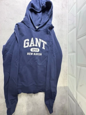 Marinblå hoodie från GANT - Snygg marinblå hoodie från GANT med tryck 'GANT 1949 NEW HAVEN' på bröstet. Klassisk känguruficka framtill och huva med dragsko. Perfekt för en avslappnad och sportig stil.