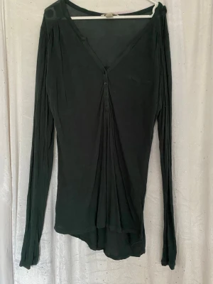 Mörkgrön blus med knappar - Mörkgrön blus med v-ringning och knappar framtill. Långärmad modell med lös passform och liten bröstficka som detalj. Tunt och mjukt material som faller snyggt. Perfekt för en avslappnad och stilren look.