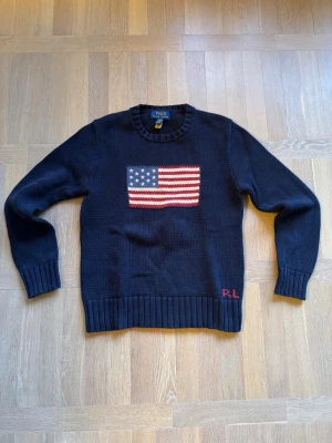 Mörkblå stickad tröja Polo Ralph Lauren - Mörkblå stickad tröja från Polo Ralph Lauren med amerikansk flagga på bröstet och RL-broderi nedtill. Nypris 2000kr+. Storlek 14/16 år