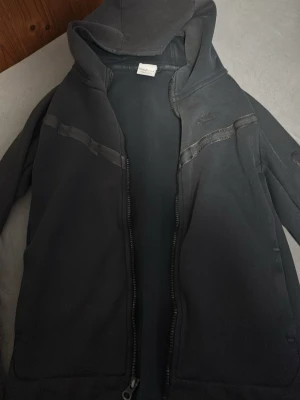 Svart hoodie från Nike med dragkedja - Säljer en svart hoodie från Nike med dragkedja framtill och huva. Hoodien har diskreta detaljer i form av svarta band över bröstet och två fickor framtill. Perfekt för en sportig och avslappnad stil.