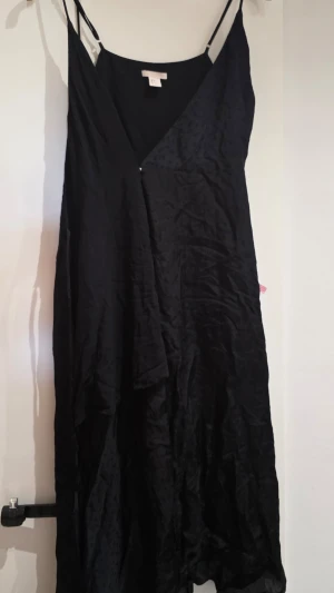 Svart omlottklänning i satin - Svart långklänning från H&M  i glansigt satinmaterial med omlott-design och tunna axelband. Klänningen har v-ringning och en elegant, flowy siluett som ger en stilren look.