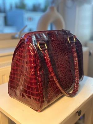 Vinröd handväska i krokopräglat skinn - Säljer en vinröd handväska med glansig krokopräglad yta och guldfärgade metalldetaljer. Väskan har två långa handtag och dragkedja upptill. Perfekt för dig som vill ha en statementväska med lyxig känsla och unik struktur.