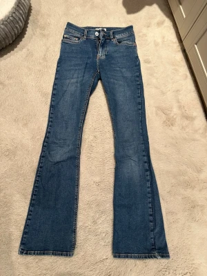 Blå bootcut jeans med mellan hög midja från Gina tricot - Snygga blå bootcut jeans med mellan hög midja och klassiska fem fickor. Jeansen har en lätt utsvängd passform nertill och stängs med knapp och dragkedja. Perfekta för dig som gillar en retroinspirerad look. Har knappt använt men fick hänga med ut i rensningen 